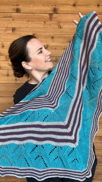 a woman is displaying a large triangle handknit shawl with alternating stripes and lace