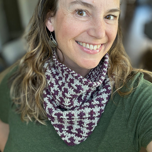 A smiling woman with light brown hair wearing a knitted cowl scarf with a purple and cream houndstooth pattern, paired with a green t-shirt in a softly blurred indoor setting.