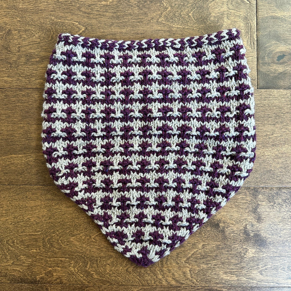 A hand-knitted cowl with a purple and cream houndstooth pattern, laid flat on a wooden floor.